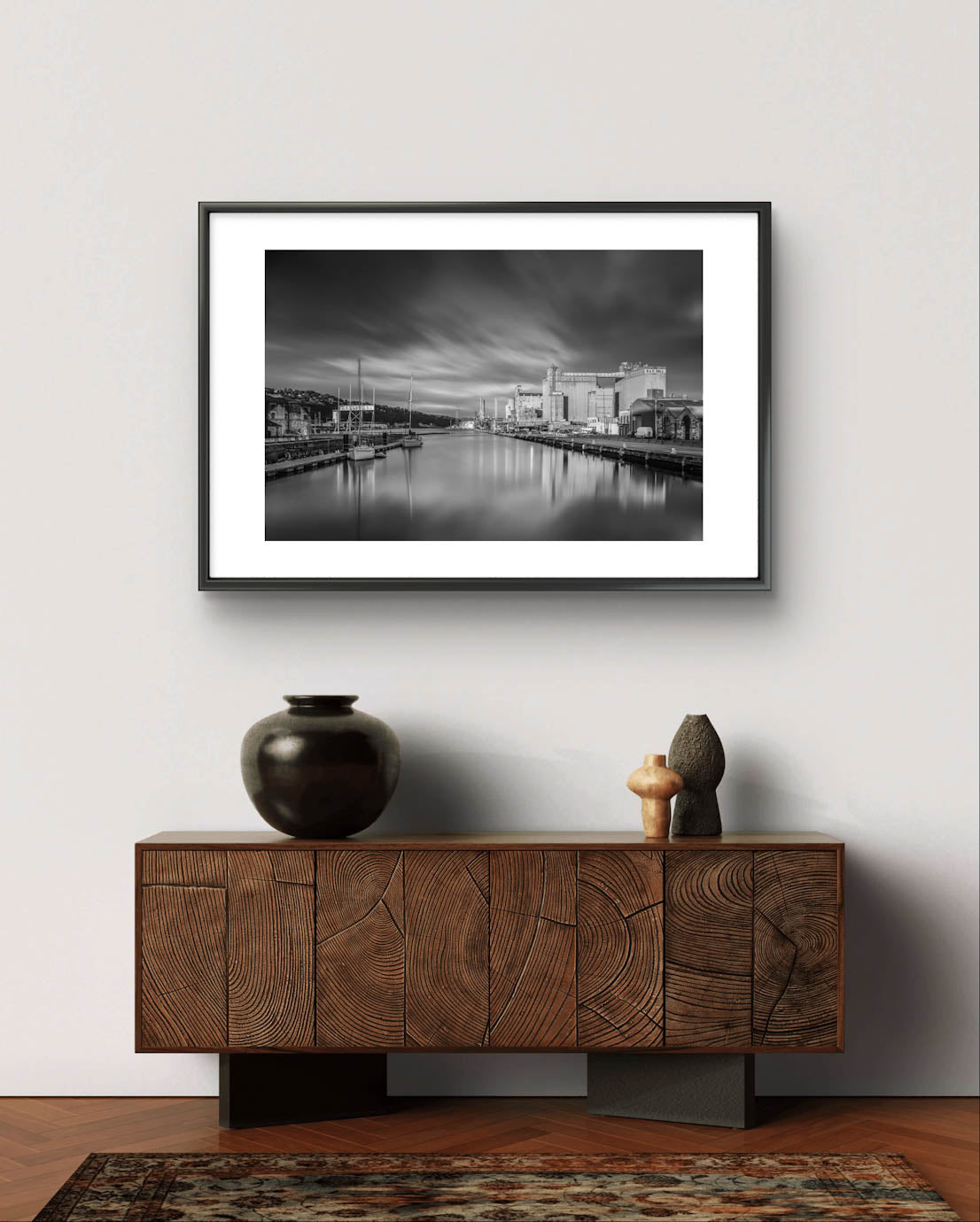 Light Grey Wall. Black and white framed photograph of a calm waterfront scene with industrial buildings along the right side and several moored boats on the left. The long exposure effect creates smooth water reflections and soft, streaked clouds in the sky, enhancing the tranquil, moody atmosphere of the port area. RH Hall, Port of Cork, Ireland. Fine Art. Limited Edition