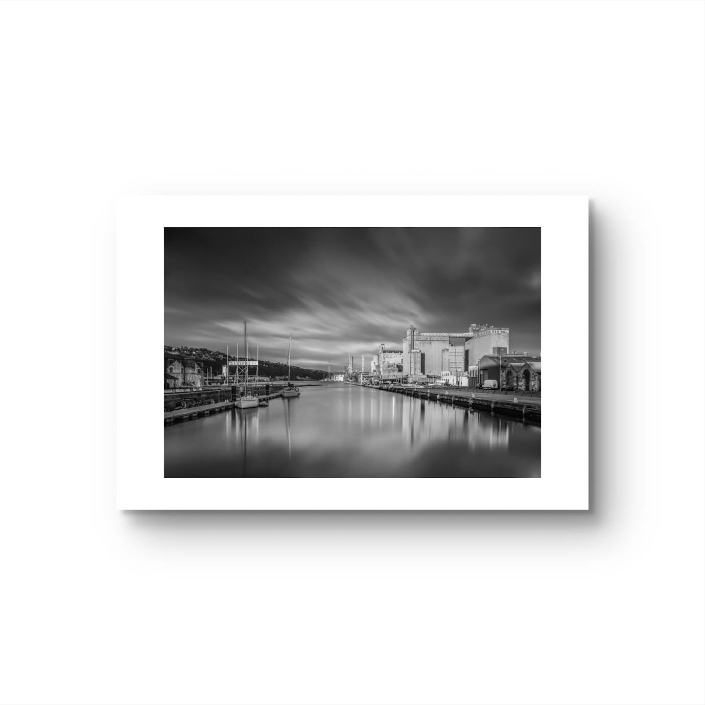 Black and white unframed photograph of a calm waterfront scene with industrial buildings along the right side and several moored boats on the left. The long exposure effect creates smooth water reflections and soft, streaked clouds in the sky, enhancing the tranquil, moody atmosphere of the port area. RH Hall, Port of Cork, Ireland. Fine Art. Limited Edition