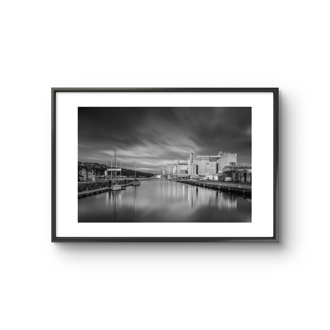 Black and white framed photograph of a calm waterfront scene with industrial buildings along the right side and several moored boats on the left. The long exposure effect creates smooth water reflections and soft, streaked clouds in the sky, enhancing the tranquil, moody atmosphere of the port area. RH Hall, Port of Cork, Ireland. Fine Art. Limited Edition