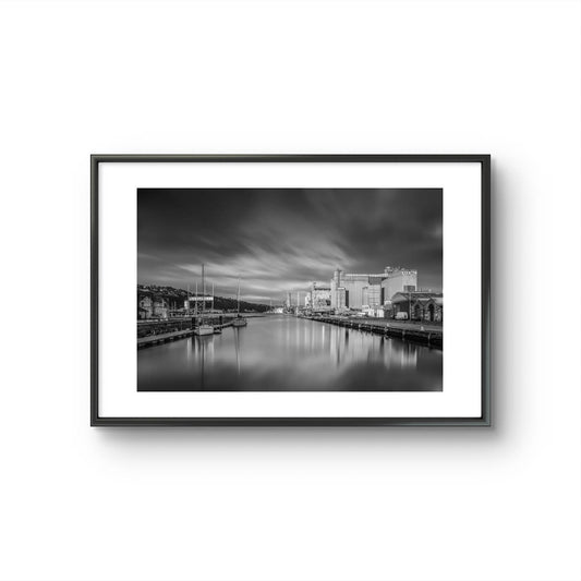 Black and white framed photograph of a calm waterfront scene with industrial buildings along the right side and several moored boats on the left. The long exposure effect creates smooth water reflections and soft, streaked clouds in the sky, enhancing the tranquil, moody atmosphere of the port area. RH Hall, Port of Cork, Ireland. Fine Art. Limited Edition