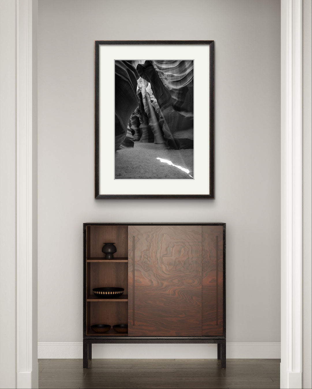 mahogany console table, grey wall and on it is Framed Fine Art black and white photograph of Canyon’s narrow passageway with the light cascading down the walls like a waterfall of illumination. The intricate patterns of the sandstone, carved by centuries of wind and water, create a mesmerizing texture that draws the eye upward. Antelope Canyon, USA, Arizona
