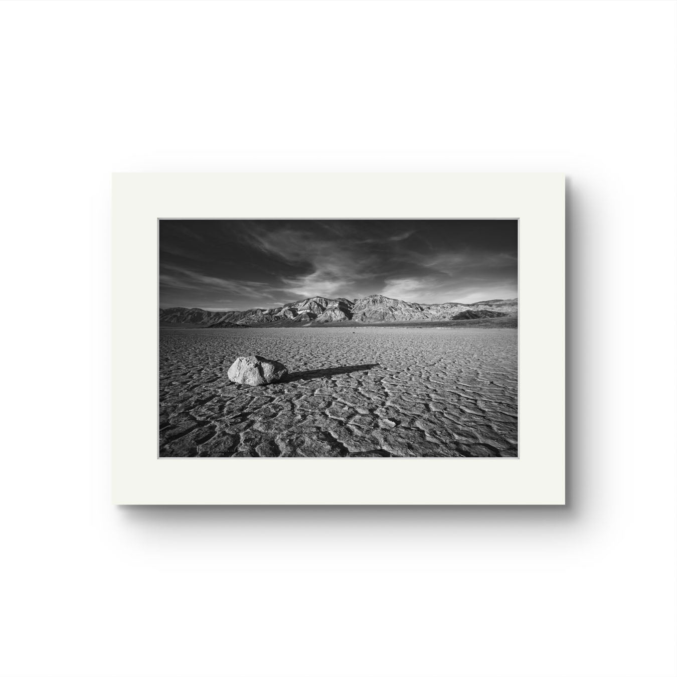 Black and white fine art photograph print of a striking desert landscape in Death Valley National Park. Featuring a solitary rock on the cracked, dry lakebed of the Racetrack Playa, this iconic scene is enhanced by dramatic skies and rugged mountain ranges in the background. Perfect for home decor, wall art, or photography enthusiasts seeking unique nature and landscape prints, this image captures the stark beauty, texture, and timeless appeal of the American Southwest.