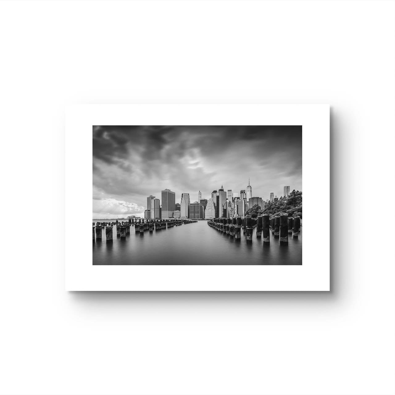 Unframed fine art, black and white photography print capturing the view of Manhattan from the Brooklyn side, across the East River. The scene is framed by old pier pillars sticking out of the water, adding intriguing leading lines that draw the viewer's gaze into the heart of the city. New York City, NYC, USA, Limited Edition Print.