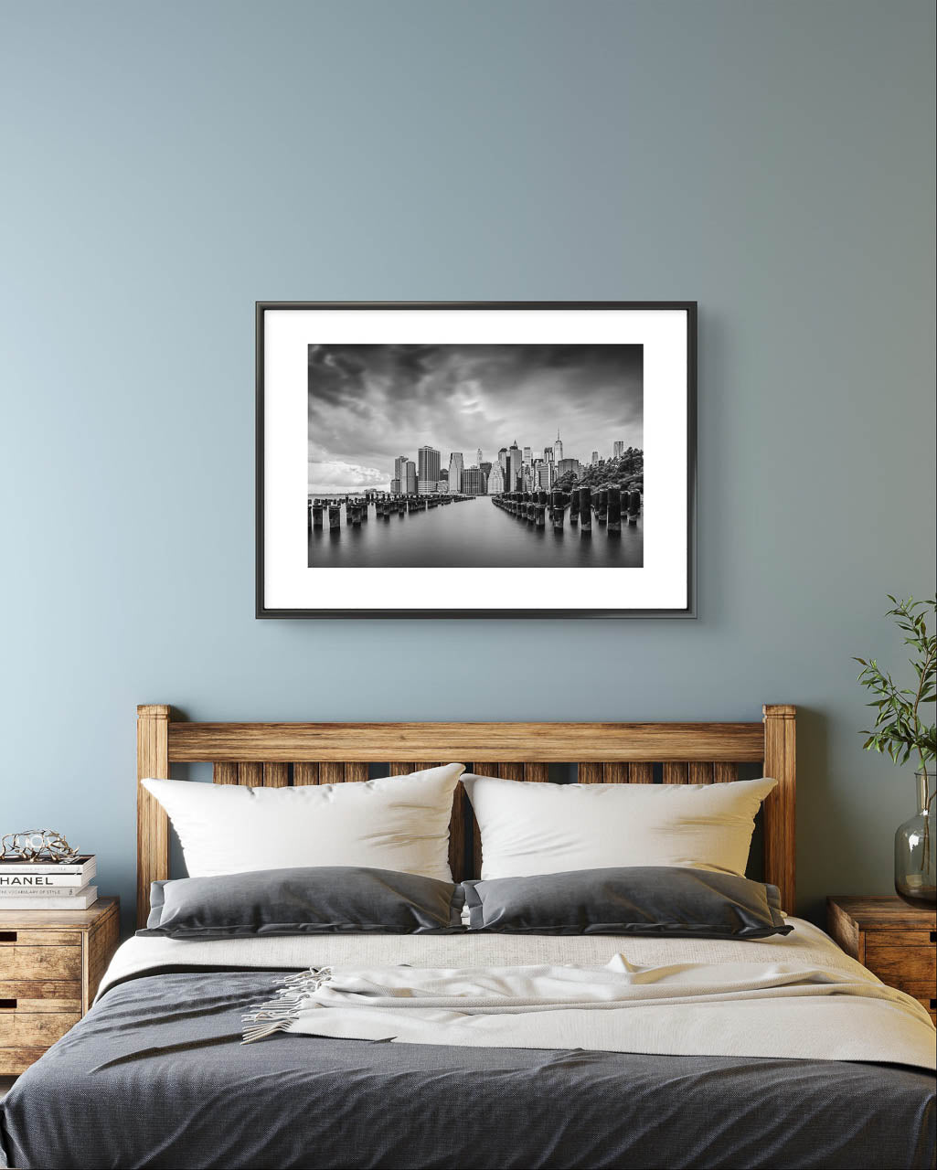 Bedroom. Large bed with bed lockers aside and above it is Framed fine art, black and white photography print capturing the view of Manhattan from the Brooklyn side, across the East River. The scene is framed by old pier pillars sticking out of the water, adding intriguing leading lines that draw the viewer's gaze into the heart of the city. New York City, NYC, USA, Limited Edition Print.