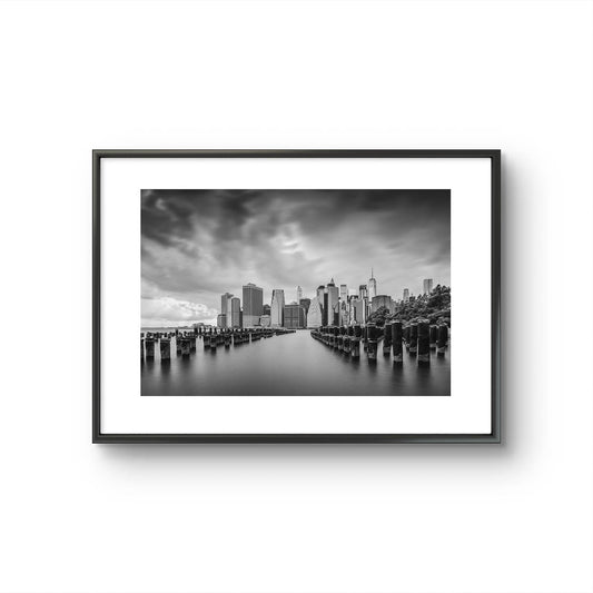 Framed fine art, black and white photography print capturing the view of Manhattan from the Brooklyn side, across the East River. The scene is framed by old pier pillars sticking out of the water, adding intriguing leading lines that draw the viewer's gaze into the heart of the city. New York City, NYC, USA, Limited Edition Print.