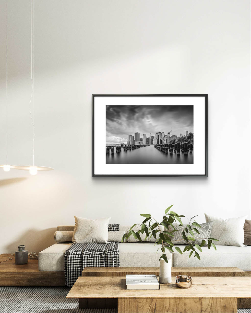 Modern home living room, on the gray wall Framed fine art, black and white photography print capturing the view of Manhattan from the Brooklyn side, across the East River. The scene is framed by old pier pillars sticking out of the water, adding intriguing leading lines that draw the viewer's gaze into the heart of the city. New York City, NYC, USA, Limited Edition Print.