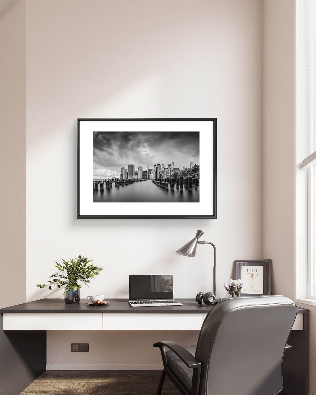 Home office on the greyish wall is Framed fine art, black and white photography print capturing the view of Manhattan from the Brooklyn side, across the East River. The scene is framed by old pier pillars sticking out of the water, adding intriguing leading lines that draw the viewer's gaze into the heart of the city. New York City, NYC, USA, Limited Edition Print.