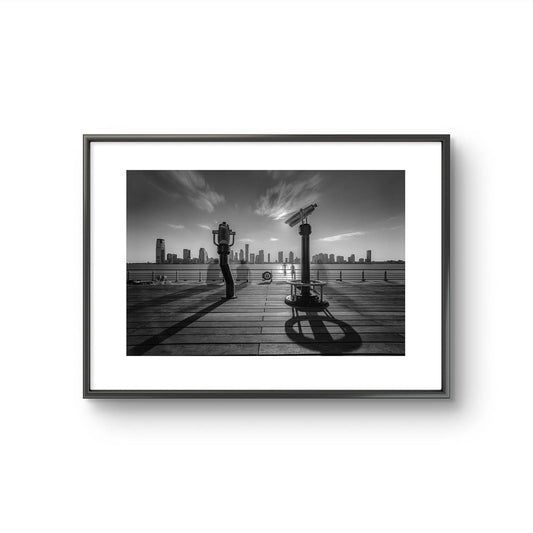 Black-and-white fine art photography print of a city skyline viewed from a waterfront deck, featuring telescopes and railings casting long shadows under a dynamic sky. Limited Edition, Pier 25 , New York City, NYC. USA