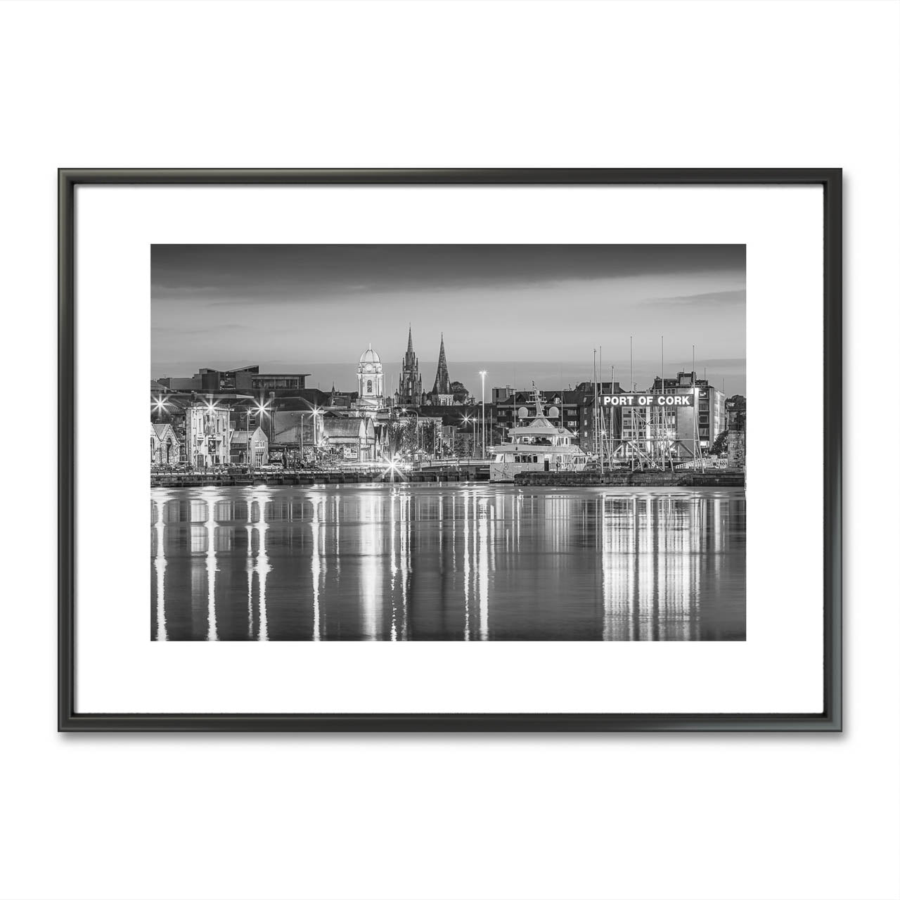 Framed black-and-white fine art photograph of Cork City, Ireland, displayed on a minimalist wall. The photo captures a serene waterfront view with reflections of historic buildings and church spires in the water. Below the artwork is a rustic wooden bench adorned with two small, textured vases, one holding a single delicate flower, adding a touch of nature and simplicity to the scene. Limited Edtion