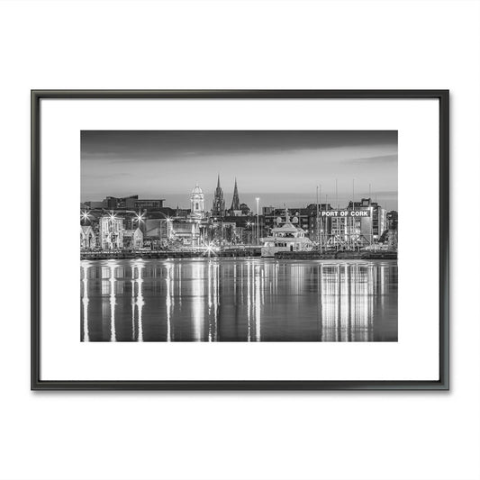 Framed black-and-white fine art photograph of Cork City, Ireland, displayed on a minimalist wall. The photo captures a serene waterfront view with reflections of historic buildings and church spires in the water. Below the artwork is a rustic wooden bench adorned with two small, textured vases, one holding a single delicate flower, adding a touch of nature and simplicity to the scene. Limited Edtion