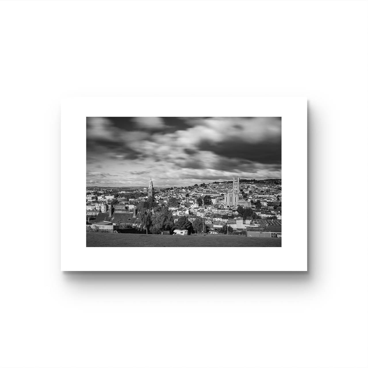 White background, Black and white photograph of a cityscape with a cloudy sky, capturing a wide view of buildings and distant hills in the background. The buildings appear detailed, with a mix of architectural styles, creating a sense of depth and texture in the urban landscape. Cork City, Ireland, Young Offenders Movie Iconic View. Bells Field. Shandon Bell's. Fine Art print, Limited Edition