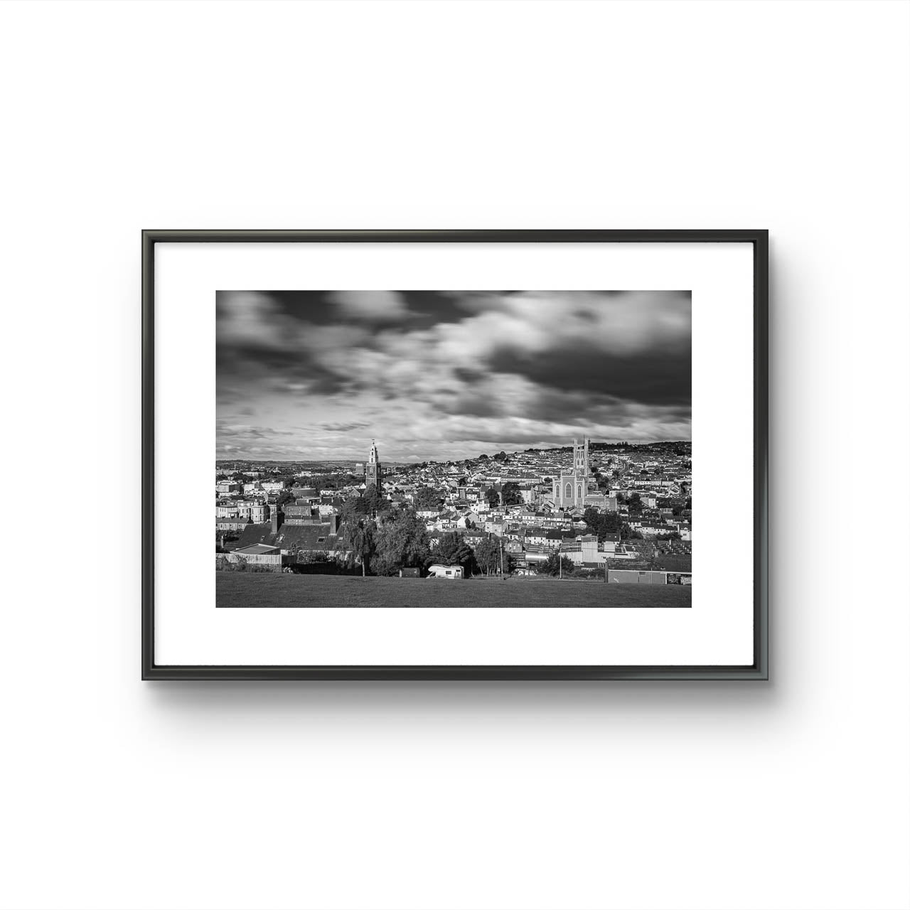 Black and white framed photograph of a cityscape with a cloudy sky, capturing a wide view of buildings and distant hills in the background. The buildings appear detailed, with a mix of architectural styles, creating a sense of depth and texture in the urban landscape. Cork City, Ireland, Young Offenders Movie Iconic View. Bells Field. Shandon Bell's. Fine Art print, Limited Edition