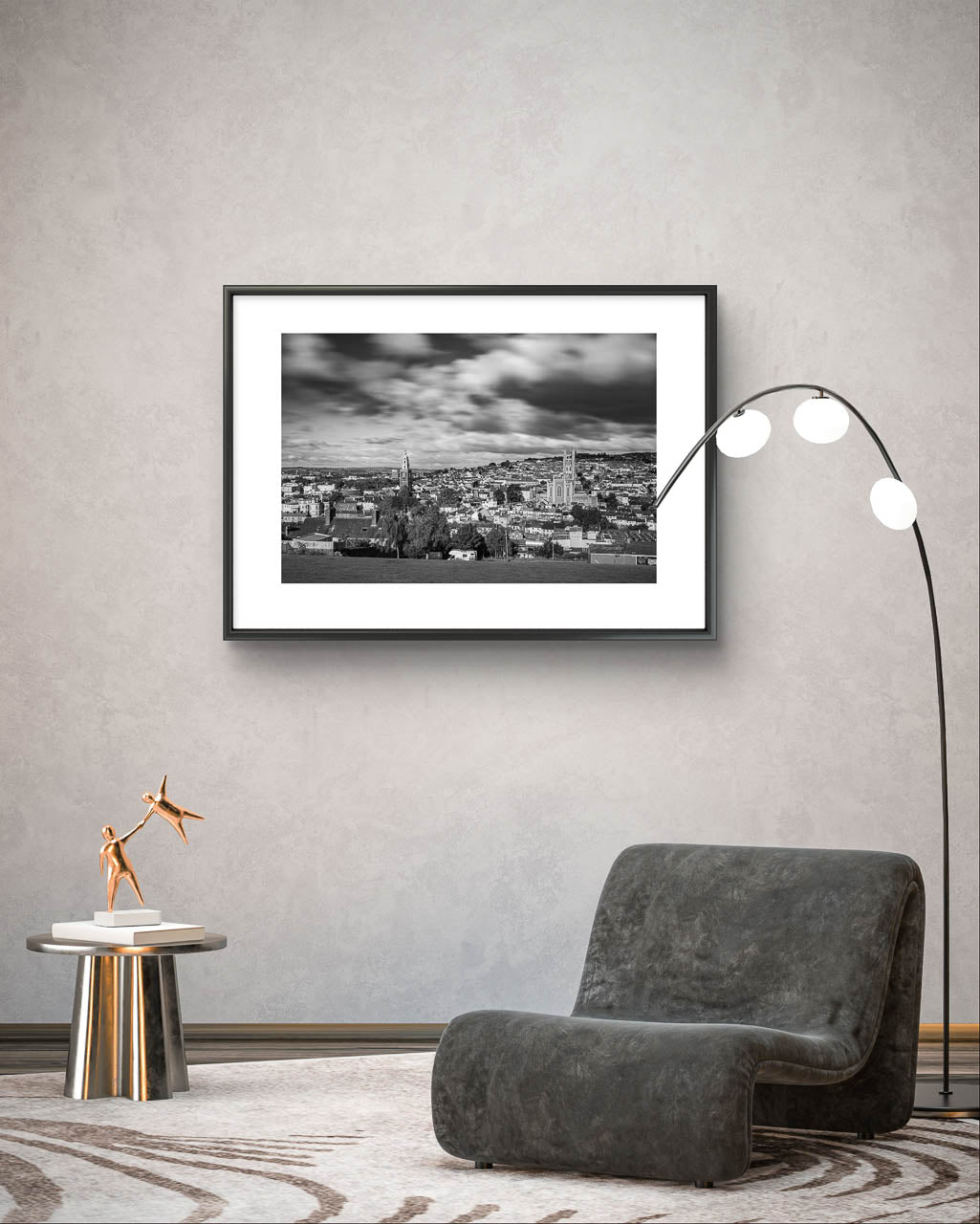 Grey Wall, Framed Black and white photograph of a cityscape with a cloudy sky, capturing a wide view of buildings and distant hills in the background. The buildings appear detailed, with a mix of architectural styles, creating a sense of depth and texture in the urban landscape. A photograph is placed in a modern room with fancy floor lamp and comfy grey chair, next it is a silver coffee table. Cork City, Ireland, Young Offenders Movie Iconic View. Bells Field. Shandon Bell's. Fine Art print Limited Edition