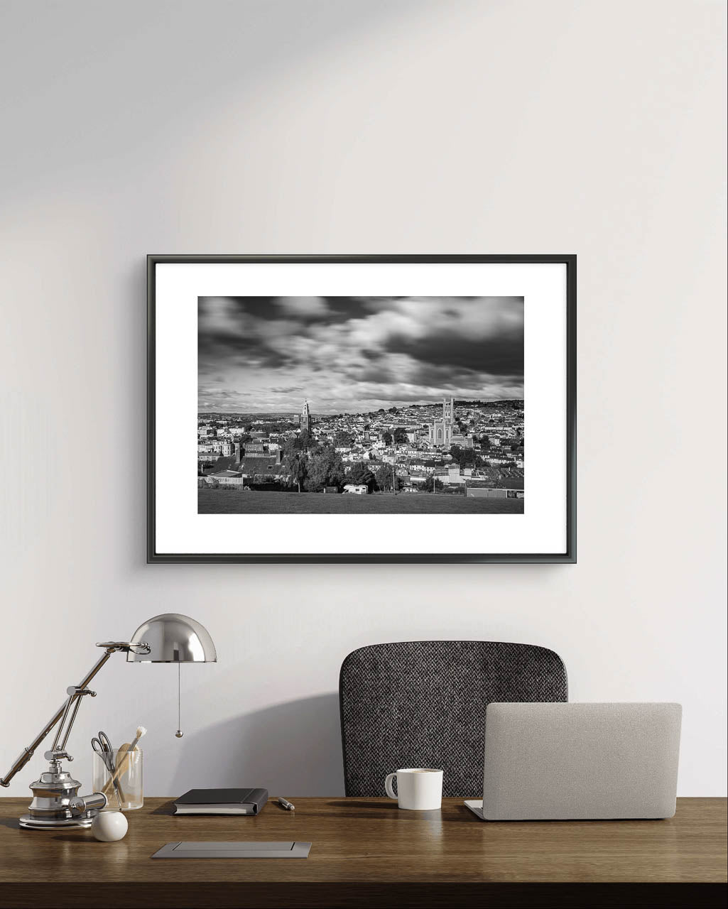 light grey wall, Black and white framed photograph of a cityscape with a cloudy sky, capturing a wide view of buildings and distant hills in the background. The buildings appear detailed, with a mix of architectural styles, creating a sense of depth and texture in the urban landscape. Picture is up over a managers desk with a laptop and coffee mug on it. Cork City, Ireland, Young Offenders Movie Iconic View. Bells Field. Shandon Bell's. Fine Art print, Limited Edition
