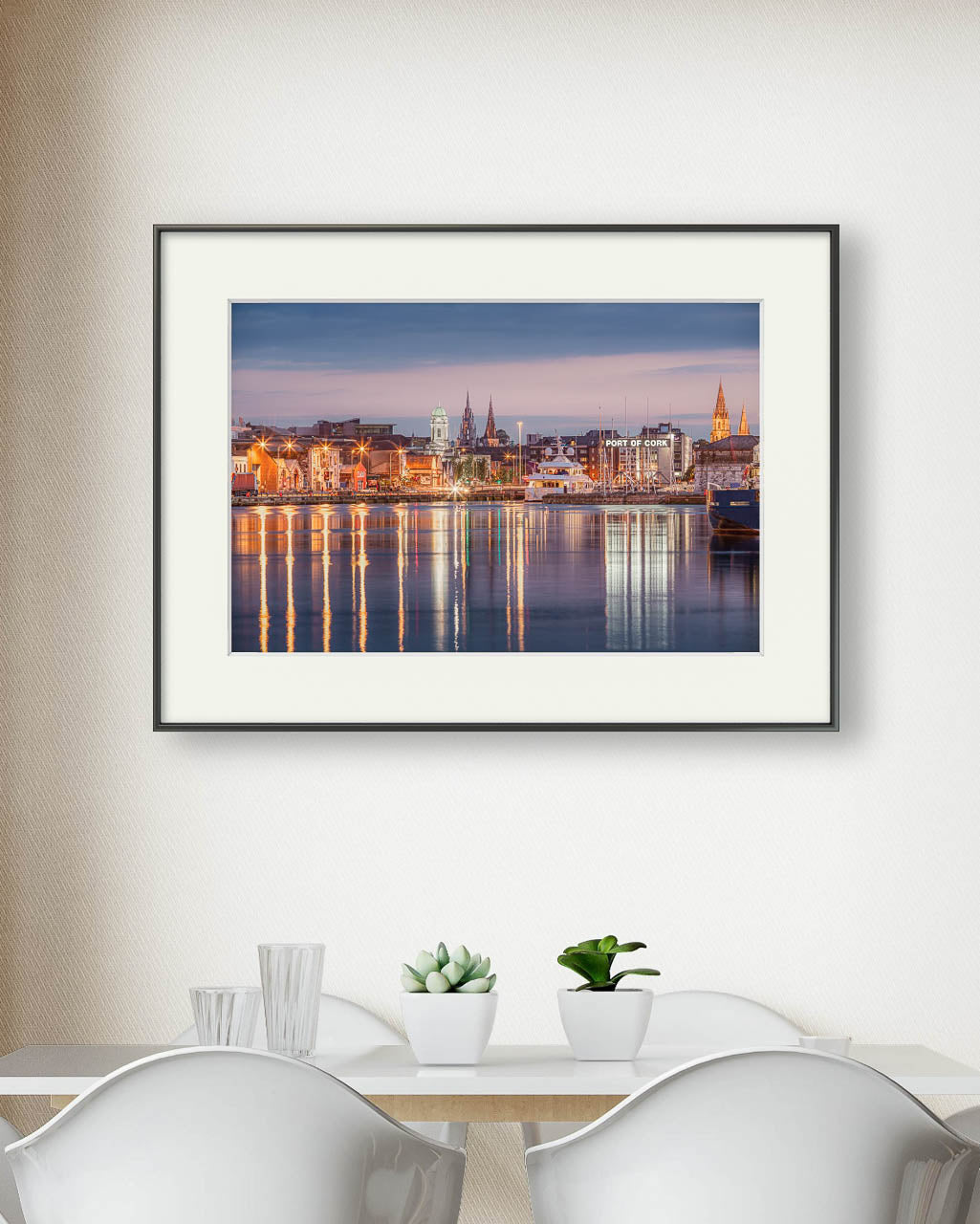 Framed colour fine art photograph of Cork, Ireland, the photo captures a serene waterfront view with reflections of historic buildings and church spires in the water. Photograph is hanged up on a white wall over the dining table.  Limited Edition print