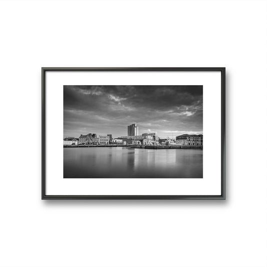 Black and white framed Fine Art 
photograph of a calm waterfront scene with industrial buildings in the focal point. The long exposure effect creates smooth water reflections. streaked clouds in the sky, enhancing the tranquil, moody atmosphere of the port area .Albert Quay & Victoria Rd. Limited Edition Print, Fine Art photography, Cork City, Ireland