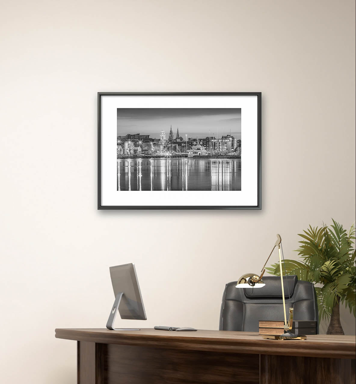 Framed black-and-white fine art photograph of Cork, Ireland, displayed on a minimalist wall. The photo captures a serene waterfront view with reflections of historic buildings and church spires in the water. Below the artwork is office desk with computer screen on, modern desk lamp and partially visible plant behind office chair.  Limited Edition