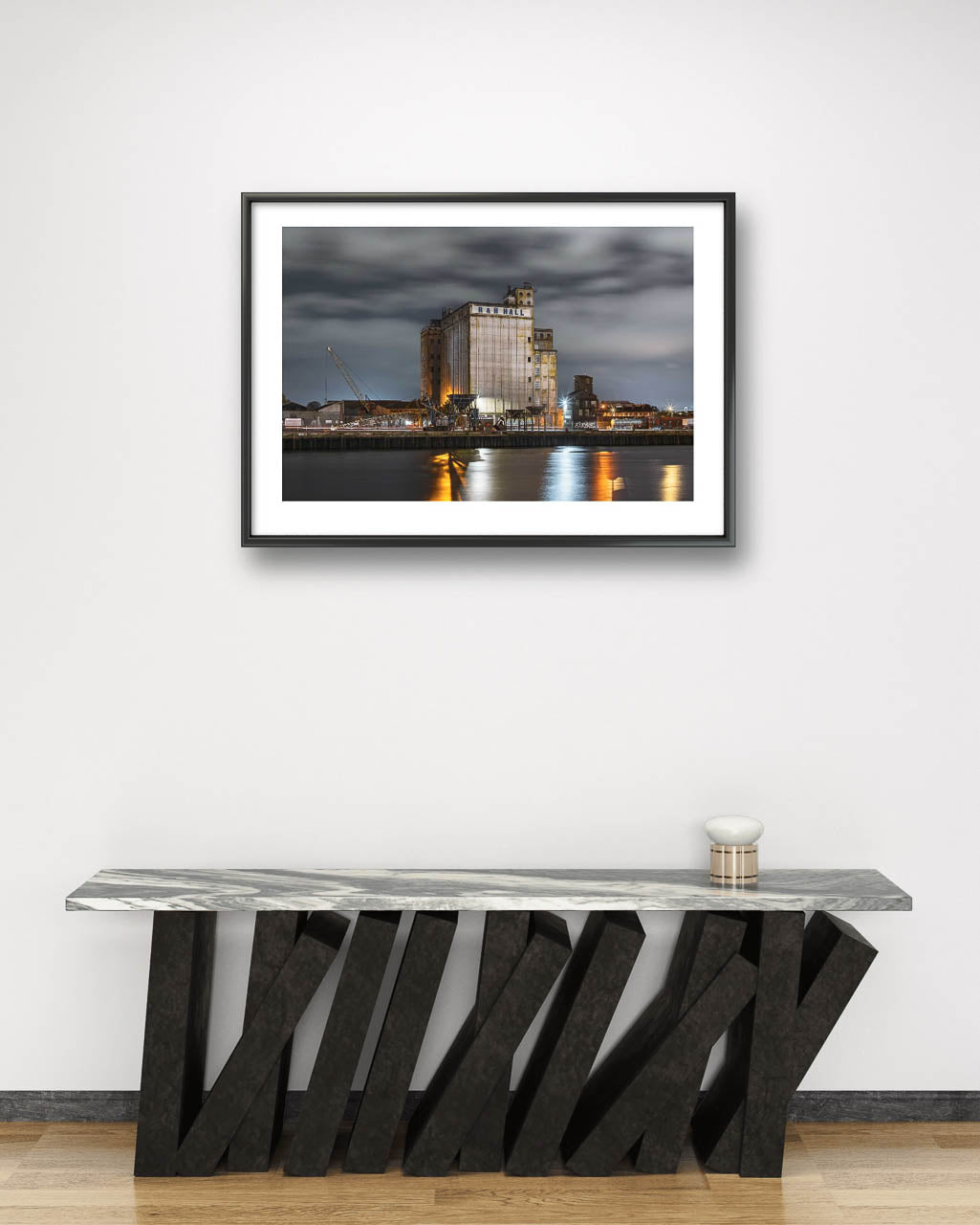 Minimalistic wall. A modern table. Framed fine art photograph of an industrial waterfront scene at night, featuring a large building labeled 'R&H Hall' with surrounding structures and cranes. The long exposure captures smooth water reflections of lights from the building, creating a calm, reflective atmosphere under a cloudy sky. Cork City, Ireland. Limited Edition Print