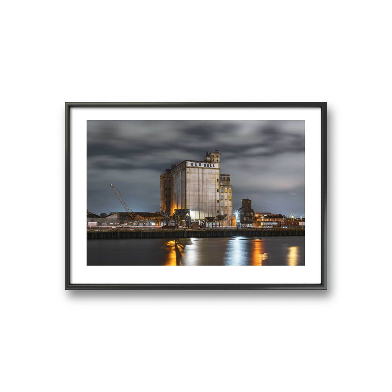 Framed fine art photography print of an industrial waterfront scene at night, featuring a large building labeled 'R&H Hall' with surrounding structures and cranes. The long exposure captures smooth water reflections of lights from the building, creating a calm, reflective atmosphere under a cloudy sky. Cork City, Ireland. Limited Edition Print