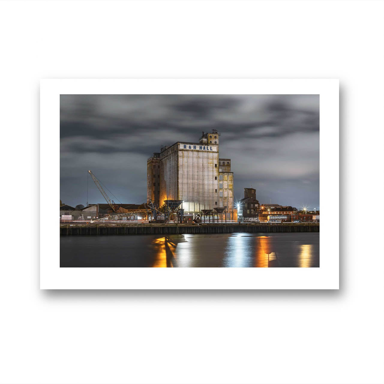 Unframed fine art photography print of an industrial waterfront scene at night, featuring a large building labeled 'R&H Hall' with surrounding structures and cranes. The long exposure captures smooth water reflections of lights from the building, creating a calm, reflective atmosphere under a cloudy sky. Cork City, Ireland. Limited Edition Print