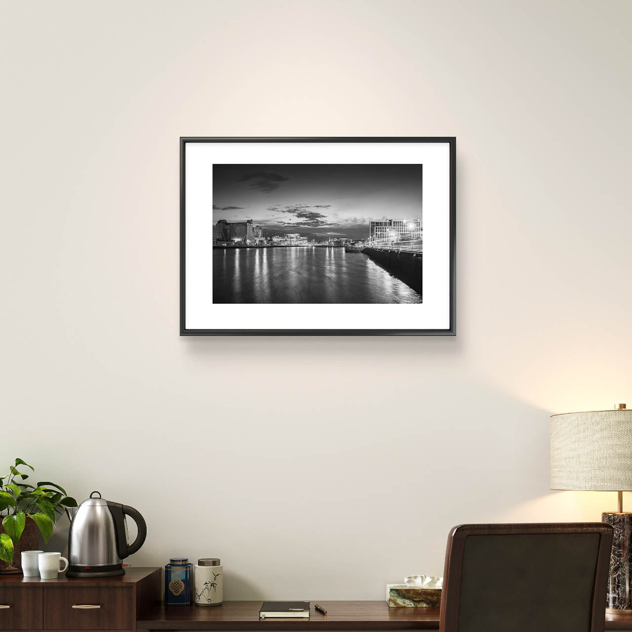 Brown desk and over it is hanging Framed Black and white photograph of a riverside cityscape at dusk. The calm river reflects lights from buildings along the shoreline, creating a shimmering effect on the water. Modern buildings line both sides of the river, with a cloudy sky adding a serene, moody atmosphere. The city lights contrast with the darkening sky, giving the scene a peaceful, contemplative quality. Cork City, Ireland