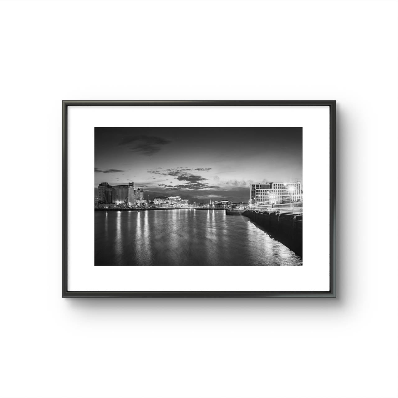 Framed Black and white Fine Art photography print of a riverside cityscape at dusk. The calm river reflects lights from buildings along the shoreline, creating a shimmering effect on the water. Modern buildings line both sides of the river, with a cloudy sky adding a serene, moody atmosphere. Horgan's Quay, Kennedy's Quay, River Lee, Cork City, Ireland. Limited Edition