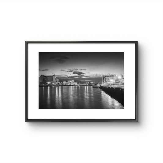 Framed Black and white Fine Art photography print of a riverside cityscape at dusk. The calm river reflects lights from buildings along the shoreline, creating a shimmering effect on the water. Modern buildings line both sides of the river, with a cloudy sky adding a serene, moody atmosphere. Horgan's Quay, Kennedy's Quay, River Lee, Cork City, Ireland. Limited Edition