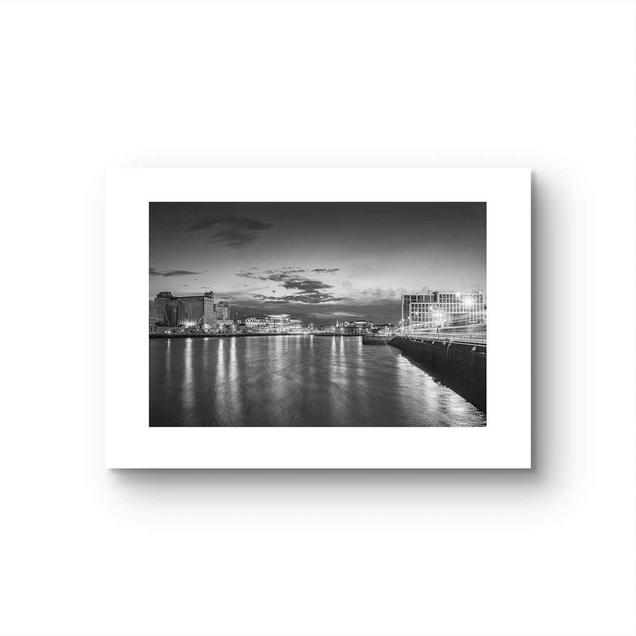 Unframed Black and white photograph of a riverside cityscape at dusk. The calm river reflects lights from buildings along the shoreline, creating a shimmering effect on the water. Modern buildings line both sides of the river, with a cloudy sky adding a serene, moody atmosphere. The city lights contrast with the darkening sky, giving the scene a peaceful, contemplative quality. Horgan's Quay, Kennedy's Quay, River Lee, Cork City, Ireland. Limited Edition, Cork City, Ireland