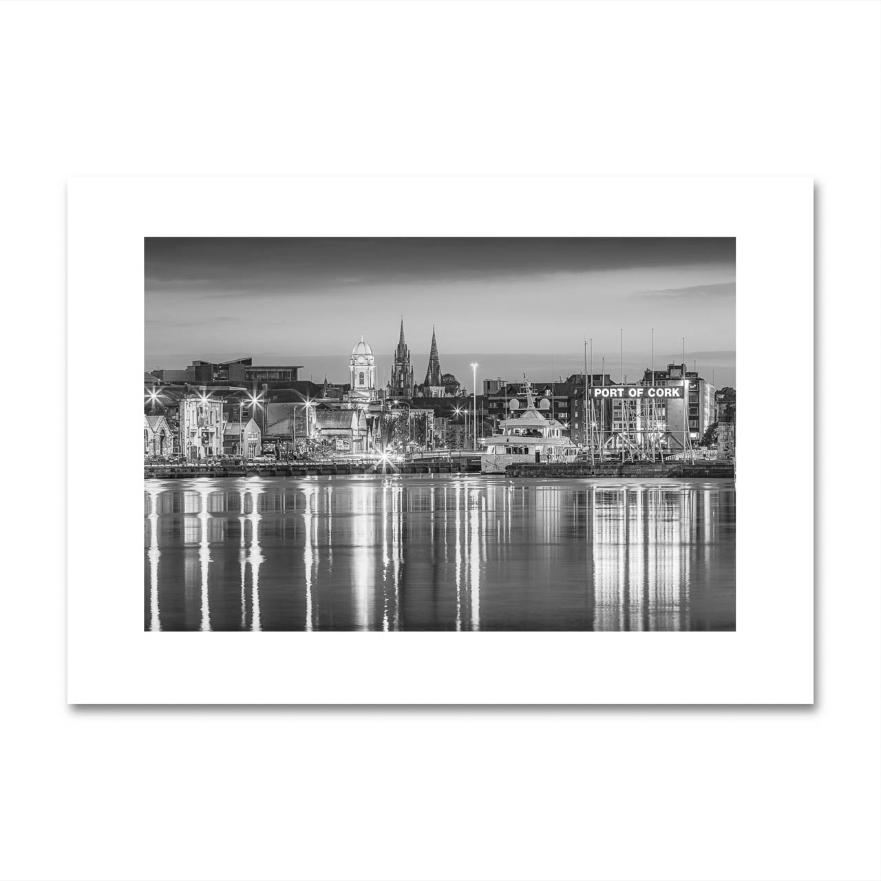 Print only of  black-and-white fine art photograph of Cork, Ireland, displayed on a minimalist wall. The photo captures a serene waterfront view with reflections of historic buildings and church spires in the water.  Limited Edition