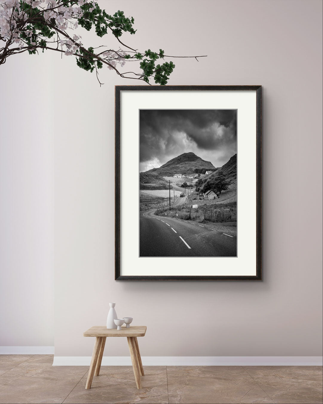 A gallery room with some plant hanging from above , small table and large Framed black and white fine art photography print showcasing Irish landscape. The twisting road and houses create a magical scenery. Ireland. Co. Donegal. Teelin