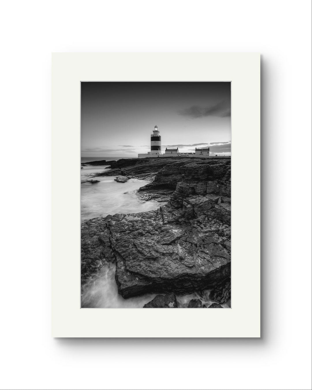 Unframed Black and white fine art photography print. This vertical composition captures the lighthouse at dusk, with the rocky coastline in the foreground leading up to the lighthouse, centered in the frame. It stands proudly against the dramatic cliffs of County Wexford. Ireland. Landscape