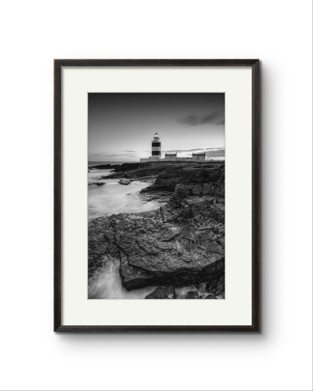 Black and white fine art photography print. Framed. This vertical composition captures the lighthouse at dusk, with the rocky coastline in the foreground leading up to the lighthouse, centered in the frame. It stands proudly against the dramatic cliffs of County Wexford. Ireland. Landscape