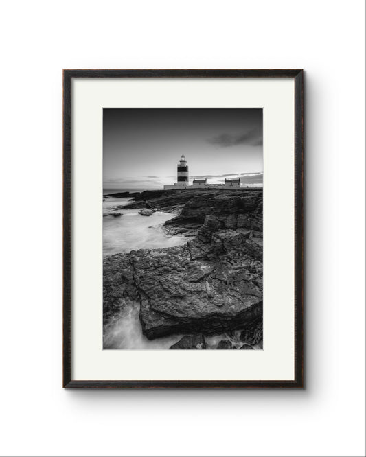 Black and white fine art photography print. Framed. This vertical composition captures the lighthouse at dusk, with the rocky coastline in the foreground leading up to the lighthouse, centered in the frame. It stands proudly against the dramatic cliffs of County Wexford. Ireland. Landscape