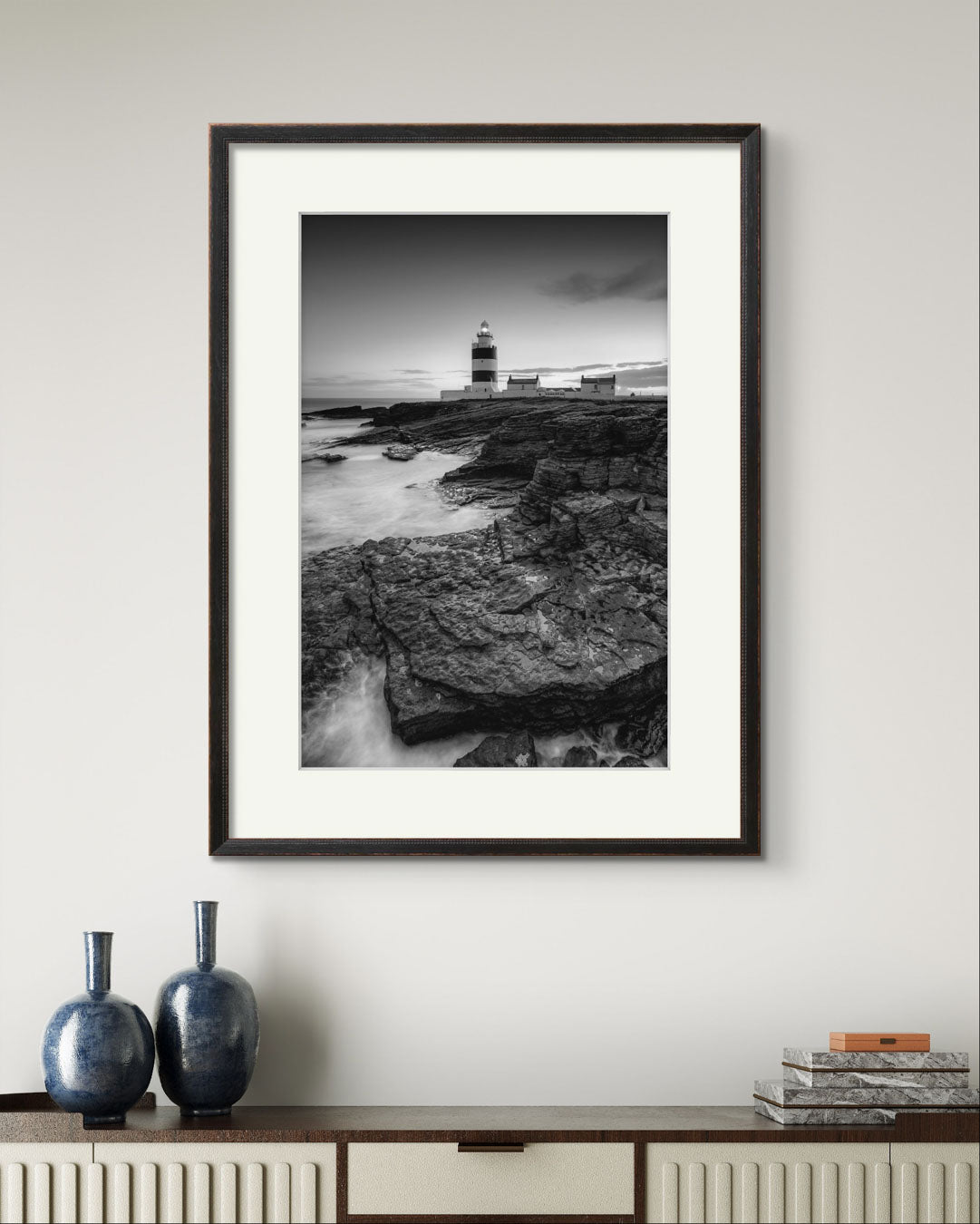 Console table with fancy vases. over it on the wall  is framed Black and white fine art photography print. This vertical composition captures the lighthouse at dusk, with the rocky coastline in the foreground leading up to the lighthouse, centered in the frame. It stands proudly against the dramatic cliffs of County Wexford. Ireland. Landscape
