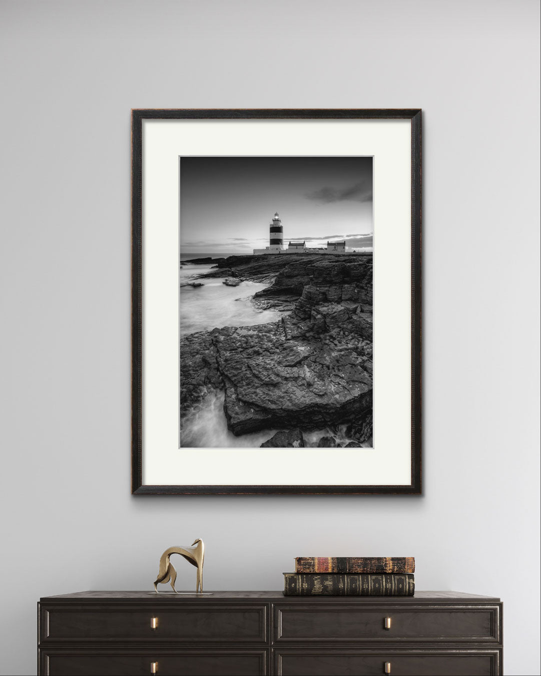 chest of drawers with some books put on top. Over it on the greyish wall is hung framed Black and white fine art photography print. This vertical composition captures the lighthouse at dusk, with the rocky coastline in the foreground leading up to the lighthouse, centered in the frame. It stands proudly against the dramatic cliffs of County Wexford. Ireland. Landscape