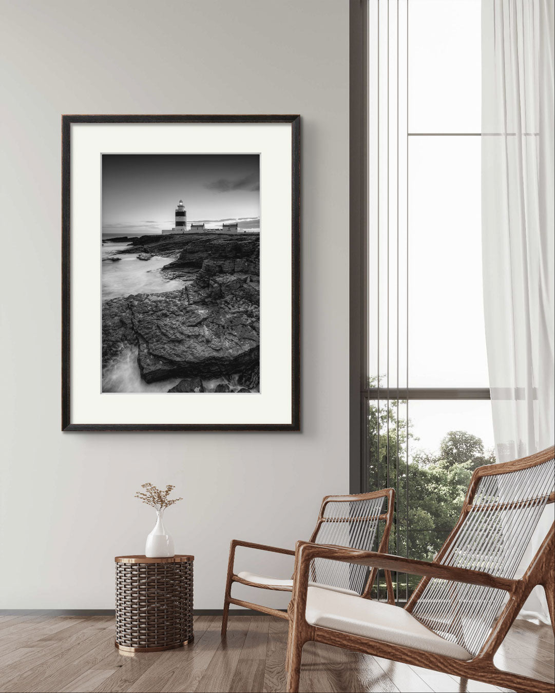 a modern sitting room . two fancy chairs and wicker basket with decorative plant. On the wall is framed Black and white fine art photography print. This vertical composition captures the lighthouse at dusk, with the rocky coastline in the foreground leading up to the lighthouse, centered in the frame. It stands proudly against the dramatic cliffs of County Wexford. Ireland. Landscape