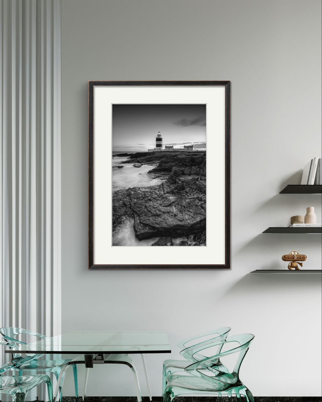A modern glass kitchen table with Plexiglas chairs. Over it framed Black and white fine art photography print. This vertical composition captures the lighthouse at dusk, with the rocky coastline in the foreground leading up to the lighthouse, centered in the frame. It stands proudly against the dramatic cliffs of County Wexford. Ireland. Landscape
