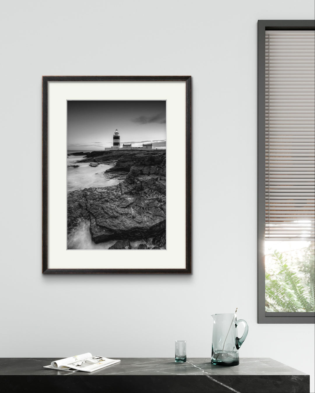 Black marble table  with glass jug on top and glass. Over it is framed Black and white fine art photography print. This vertical composition captures the lighthouse at dusk, with the rocky coastline in the foreground leading up to the lighthouse, centered in the frame. It stands proudly against the dramatic cliffs of County Wexford. Ireland. Landscape
