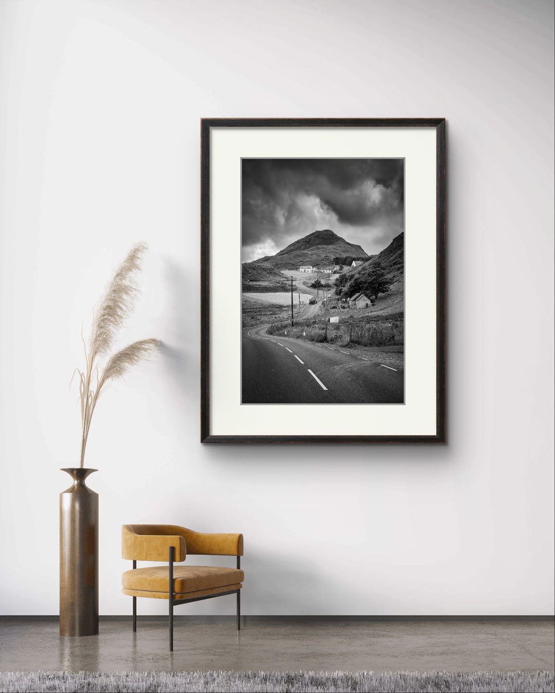 A waiting room. A chair and a decorative plant. on the wall is Framed black and white fine art photography print showcasing Irish landscape. The twisting road and houses create a magical scenery of Rural Ireland. Co. Donegal. Teelin