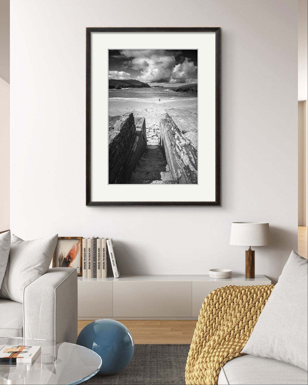 A living room and on the back wall framed photograph of an Irish Coast, dynamic composition draws your eyes to an ocean and a person walking on the distance beach. Clogher Beach. Dingle Town. Slea Head Drive in County Kerry.