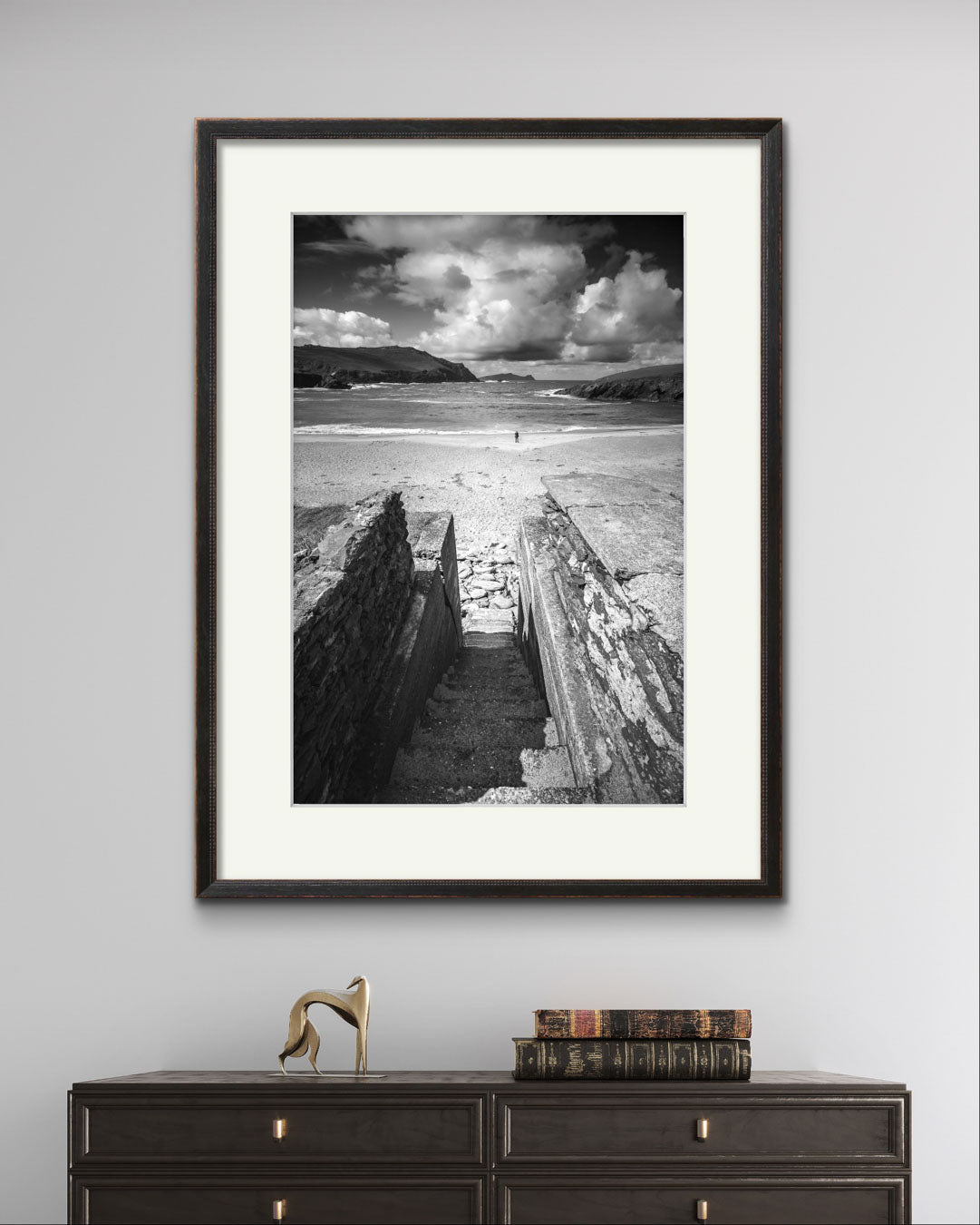 a console table with books on top. above is framed photograph of an Irish Coast, dynamic composition draws your eyes to an ocean and a person walking on the distance beach. Clogher Beach. Dingle Town. Slea Head Drive in County Kerry.