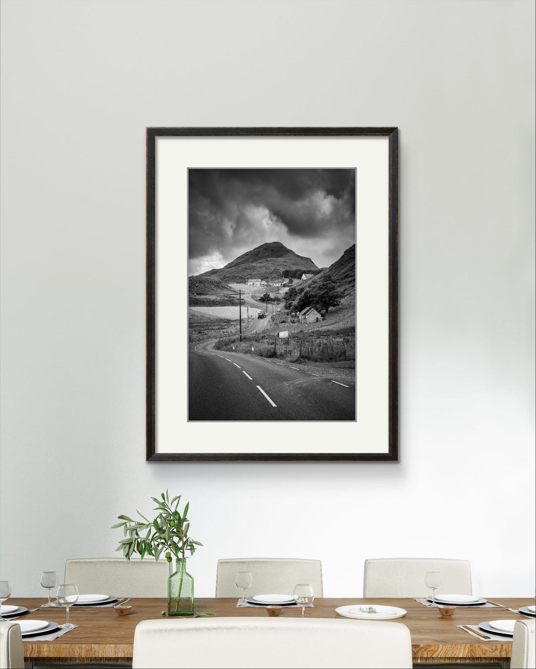 A dining room, a table with plates and glasses ready to put food on it. on the wall Framed black and white fine art photography print showcasing Irish landscape. The twisting road and houses create a magical scenery. Ireland. Co. Donegal. Teelin