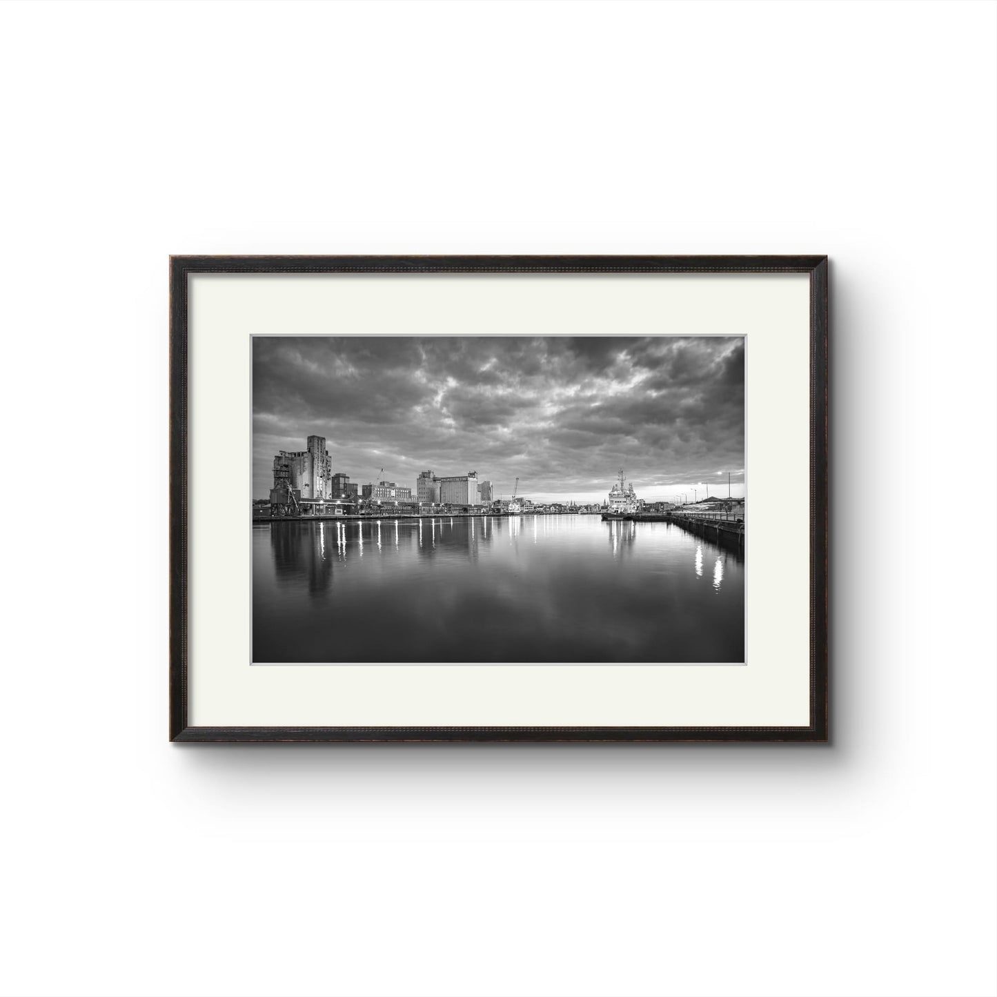 Black and white Fine Art Photography Print. The calm river reflects lights from buildings along the shoreline, creating a shimmering effect on the water. A cloudy sky adding a serene, moody atmosphere. The industrial buildings lights contrast with the darkening sky, giving the scene a peaceful atmosphere. RH Hall, Kennedy Quay, Port of Cork, Limited Edition Print, Cork City, Ireland