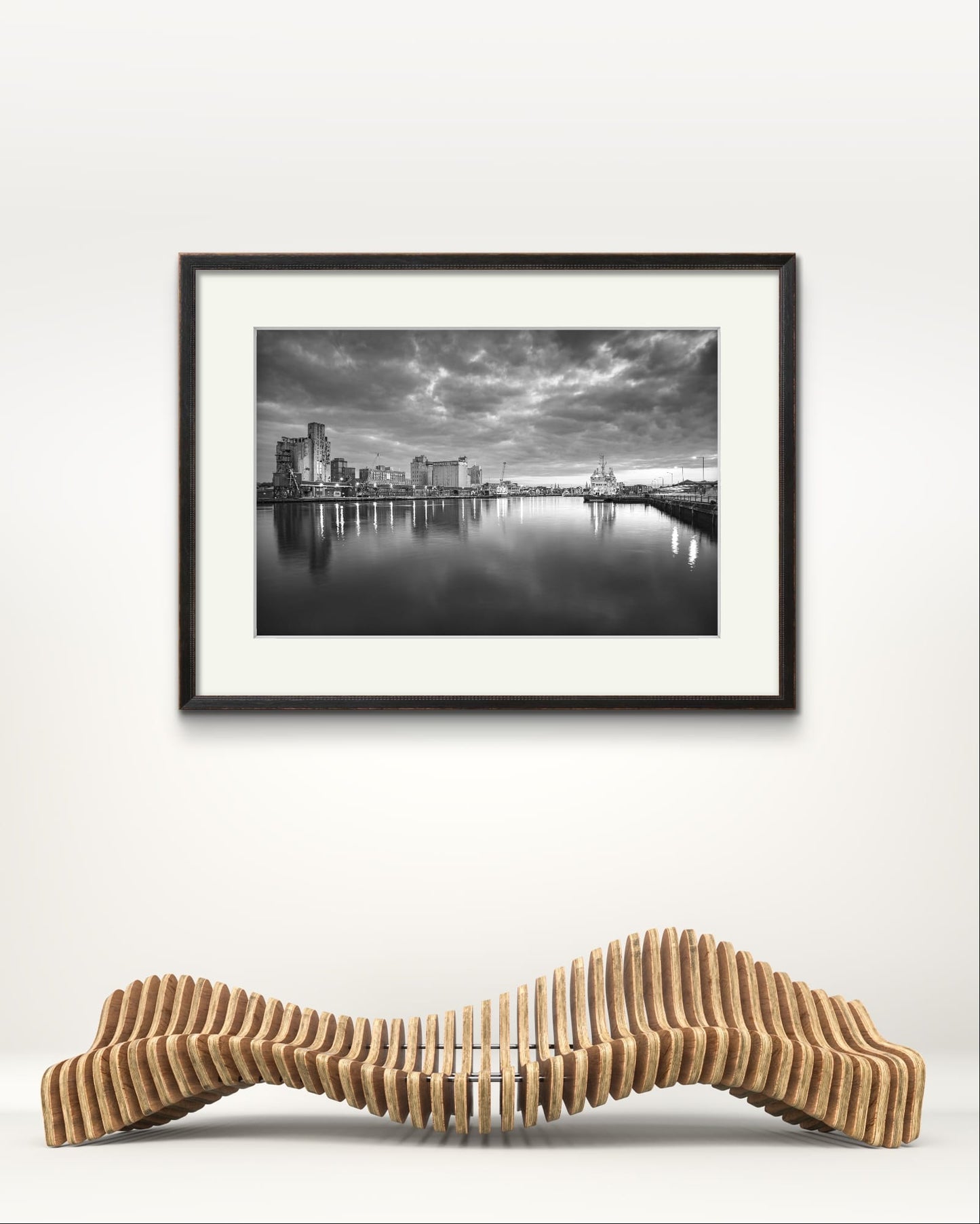 Gallery bench and Black and white Fine Art, framed photograph of a riverside. The calm river reflects lights from buildings along the shoreline, creating a shimmering effect on the water. A cloudy sky adding a serene, moody atmosphere. Kennedy Quay, Port of Cork, Limited Edition Print, RH Hall, Cork City, Ireland