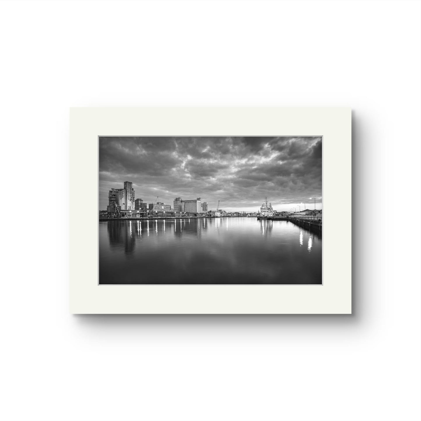 Unframed Black and white Fine Art photograph of a riverside. The calm river reflects lights from buildings along the shoreline, creating a shimmering effect on the water. A cloudy sky adding a serene, moody atmosphere. RH Hall, Kennedy Quay, Port of Cork, Limited Edition Print, Cork City, Ireland