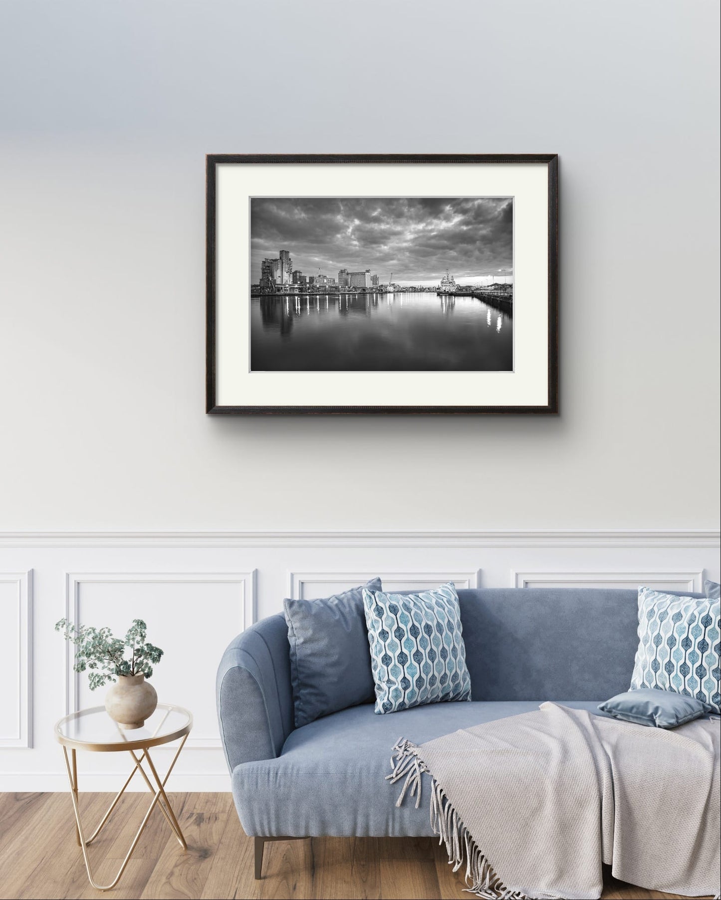 Light blue sofa with a throw and cushions. Over it is Black and white Fine Art photography Print. The calm river reflects lights from buildings along the shoreline, creating a shimmering effect on the water. A cloudy sky adding a serene, moody atmosphere. The industrial buildings lights contrast with the darkening sky, giving the scene a peaceful atmosphere. Kennedy Quay, Port of Cork, Limited Edition Print, RH Hall, Cork City, Ireland