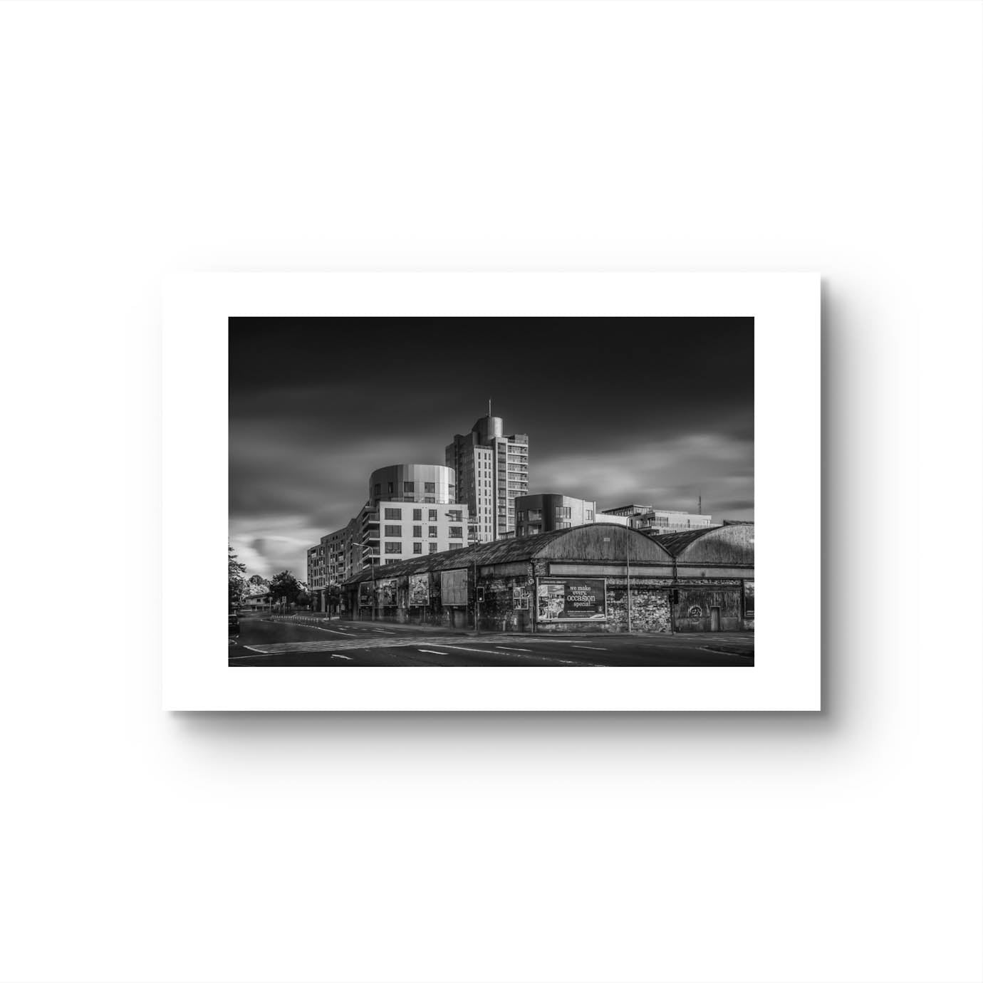 Black and white photograph of an urban street corner with a row of old, weathered warehouses in the foreground. The warehouses are covered with vintage posters and graffiti. In the background, modern high-rise buildings stand against a dramatic, cloudy sky, contrasting the historic architecture in the foreground. The photo has a moody, timeless atmosphere. Cork City, Ireland. Fine Art Photography. Limited Edition.