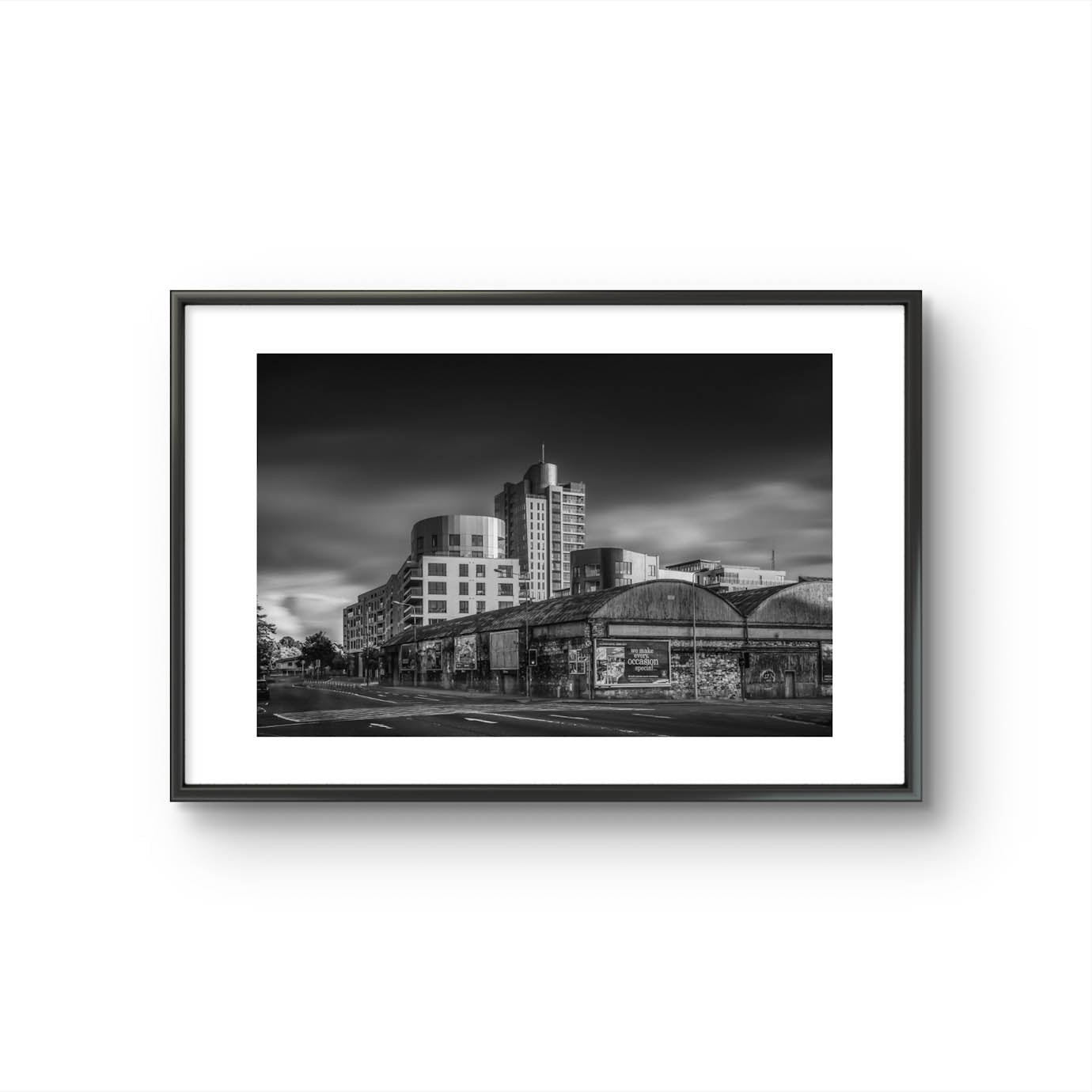Framed Black and white photograph of an urban street corner with a row of old, weathered warehouses in the foreground. The warehouses are covered with vintage posters and graffiti. In the background, modern high-rise buildings stand against a dramatic, cloudy sky, contrasting the historic architecture in the foreground. The photo has a moody, timeless atmosphere. Cork City, Ireland. Fine Art Photography. Limited Edition.
