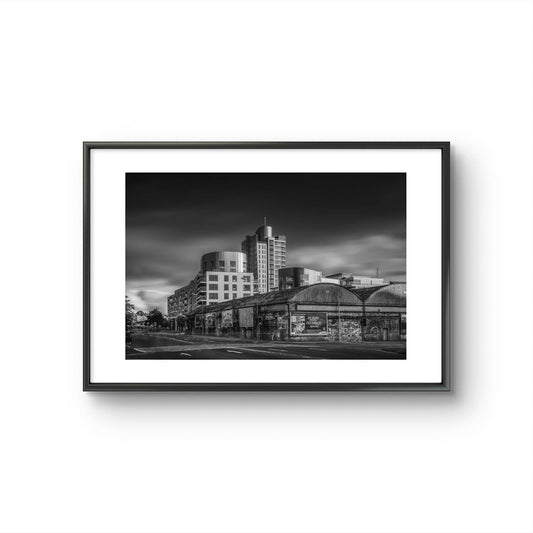 Framed Black and white photograph of an urban street corner with a row of old, weathered warehouses in the foreground. The warehouses are covered with vintage posters and graffiti. In the background, modern high-rise buildings stand against a dramatic, cloudy sky, contrasting the historic architecture in the foreground. The photo has a moody, timeless atmosphere. Cork City, Ireland. Fine Art Photography. Limited Edition.