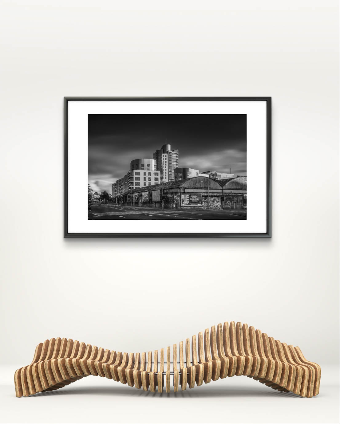 Waved gallery bench, on the wall framed Black and white photograph of an urban street corner with a row of old, weathered warehouses in the foreground. The warehouses are covered with vintage posters and graffiti. In the background, modern high-rise buildings stand against a dramatic, cloudy sky, contrasting the historic architecture in the foreground. The photo has a moody, timeless atmosphere. Cork City, Ireland. Fine Art Photography. Limited Edition.